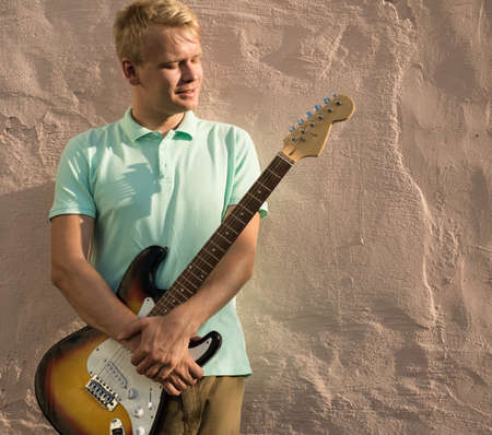 Stylish young blond man with a guitar standing against the wall, copy space.