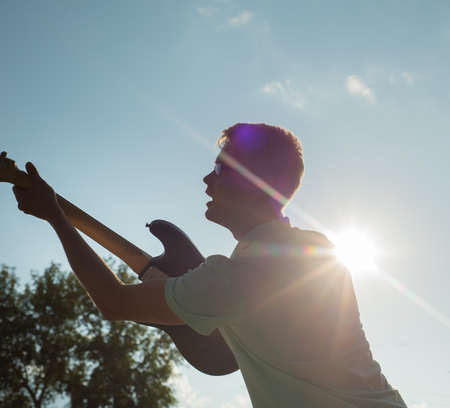 young man standing with a guitar on background of sky and sunlight.の写真素材