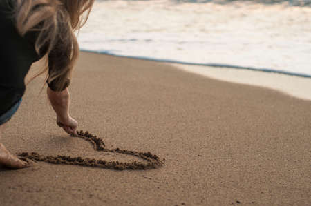 young girl draws heart on sand on sea background.の写真素材
