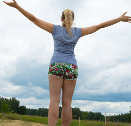 Happy young blond woman, dressed in shorts and a t-shirt stands on the road against the sky arms outstretched.の写真素材