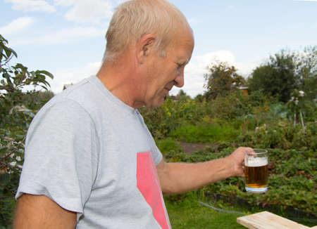An elderly man with delight and pleasure is drinking cold beer from a mug on a background of trees in your garden.の写真素材