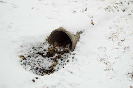 Cement sewer drain pipe, a hole with leaves and dirt, covered with the first snowの写真素材