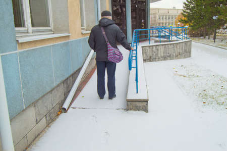 An elderly gray-haired sick man climbs hard on a snow-covered ramp for the disabled, leaving traces in the snowの写真素材