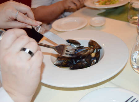 Elegant womens hands neatly cut mussels in shells, with a fork and a knife, the background of the...