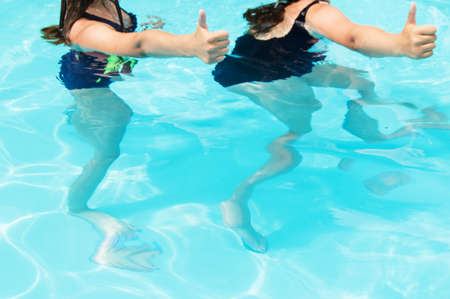 Thumbs up two teenage girls with long hair in swimsuits in a pool with blue sparkling water in the...