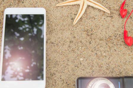 Beach vacation and travel concept, smartphone, camera, headphones and starfish on the sand on the beach, sunlight reflected in the smartphone screen.の写真素材