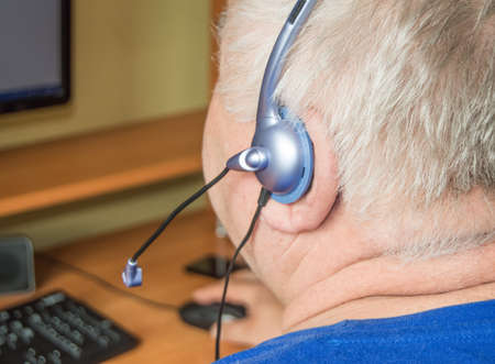 A gray-haired fat old man with a headset looks at the monitor screen, remote work, computer...