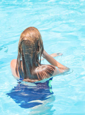 Happy girl with wet hair standing in the water in an outdoor pool, having fun in the water on vacation, vertical photo, back viewの写真素材