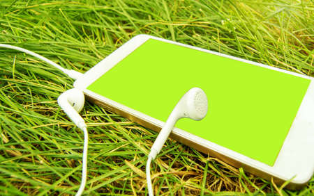 Headphones and a White phone on the green grass. Chromakey green screen of a mobile phone. Banner template with text space.の写真素材