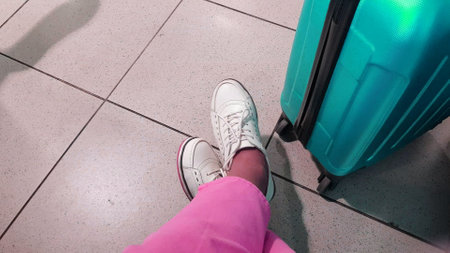 Close-up of a woman's legs in pink trousers and white sneakers, next to a blue suitcase, a woman is waiting for her plane flight at the departure terminal. The concept of travel and recreation.の写真素材