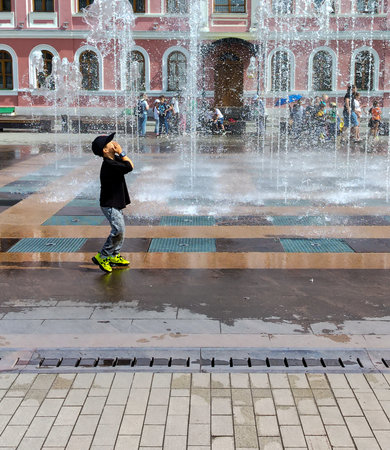 One little boy in black cap runs down the street and plays with the jets of water from a fountain spurting out of the ground.の写真素材