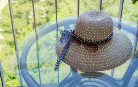 Summer straw hat and sunglasses close-up on a glass table on the balcony.の写真素材
