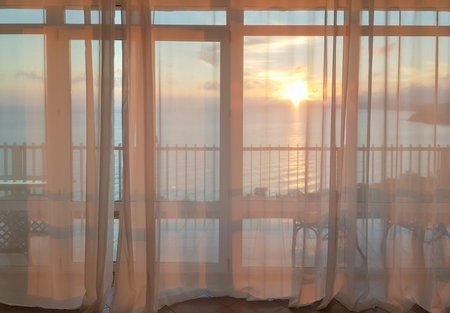 Sunset over the sea through sheer curtains at a balcony panoramic window, travel romance, meditative moodの写真素材