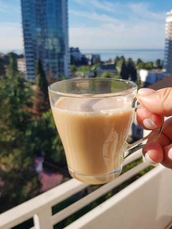A glass mug of latte held by a hand on a sunny balcony. Blurred cityscape and a sea line in the background, vertical composition, natural light, cozy mood, soft focus, shallow depth of fieldの写真素材