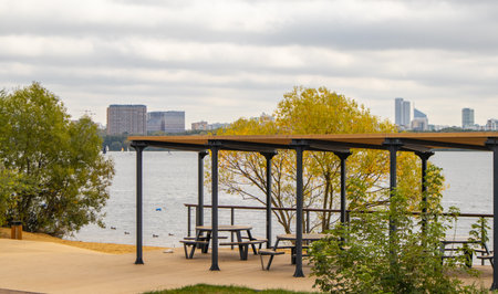 Panoramic view of Stroginsky Bay in Moscow with a pergola and picnic tables on a deck by the water. Residential skyline and small sailboats on a cloudy day. Wide copy space for design. sustainable tourism, urban nature, city break, weekend getaway,の写真素材
