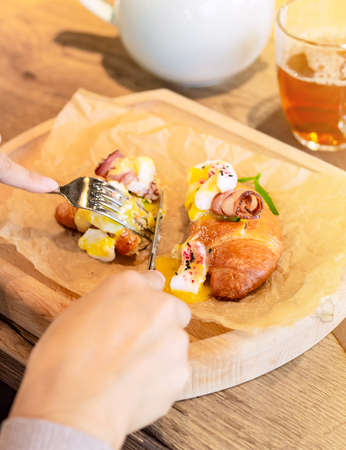 Woman has breakfast in a restaurant, egg benedict with croissant on a wooden plate under baking paper, brunch food styleの写真素材