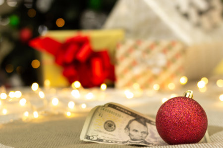 Red shiny christmas decoration with rolled-up US 5 dollar bill, gifts with bright lights and xmas tree on blurred background, festive greeting card, selective focusの写真素材