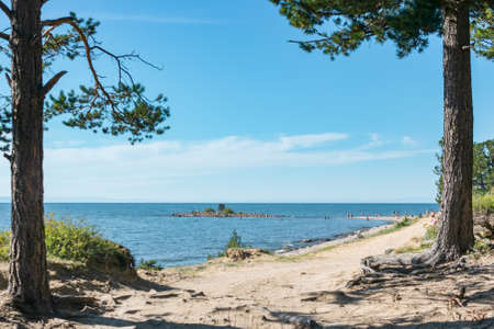 Ground back road along coast of blue sea with little island at summer sunny day, beautiful scenic desert landscape of walking vacation on lake Baikal, shore stroll away, natural frame by pine treesの写真素材