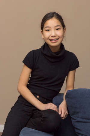 Cute female portrait of smiling teenager, young pretty asian teenage girl in dark clothes on blue couch at home looking at camera, vertical imageの写真素材