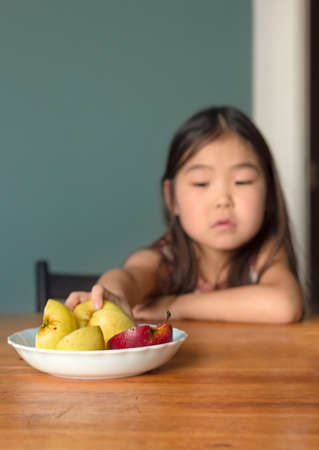 Little asian girl child takes apple fruit in kitchen at home, apartment lifestyle, healthy food concept, depth of field, front selective focus, vertical imageの写真素材
