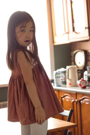 Portrait of cute little asian girl child in kitchen at home in backlight, looking at camera, apartment life, domestic lifestyle, vertical imageの写真素材