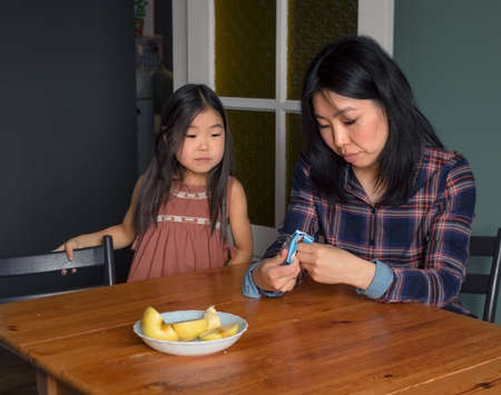 Candid asian family mom and daughter in kitchen at home, mother gives treat to girl child at table, cozy apartment life, domestic lifestyleの写真素材