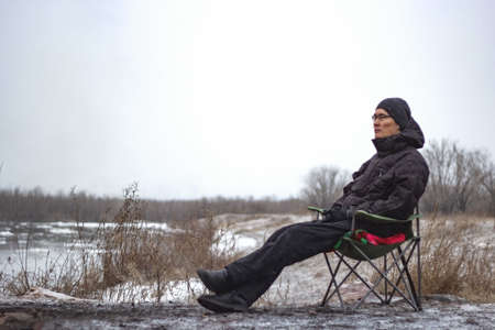 Resting brooding asian man sit on camping chair by frosty river in november, cold cloudy overcast...