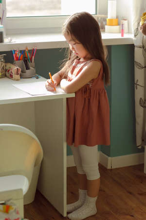 Little adorable serious asian girl child draws on table standing by window in nursery, home study...
