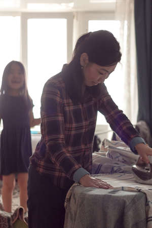 Asian family mom and daughter irons at home in living room, routine housekeeping and ironing in apartment, candid moments of domestic cleaning household together, authentic lifestyle, vertical imageの写真素材