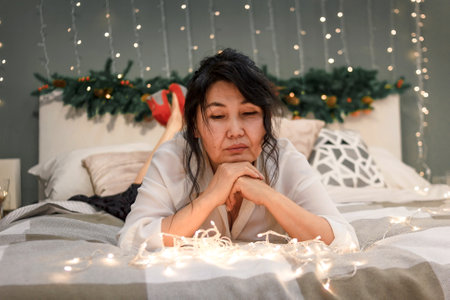 Senior asian woman relax on bed looking at xmas lights, portrait of attractive elderly sad lady in bedroom at home at christmas holidayの写真素材