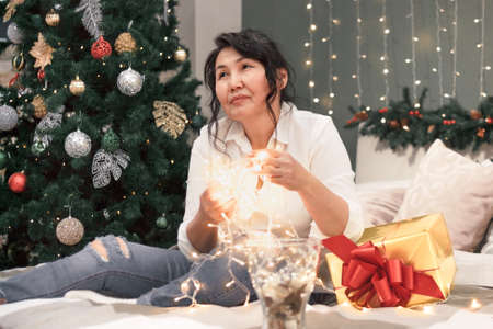 Senior asian woman on bed with xmas lights, portrait of attractive elderly lady with garland in hands and gold gift at home at christmas holidayの写真素材
