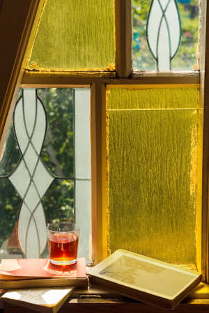 Old books and glass drink on vintage window with stained glasses and mosaic in country home of...