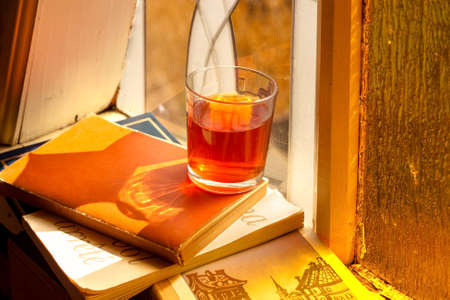 Old books and glass drink on vintage window with stained glasses and mosaic in country home of countryside, cozy still life of rustic mood with hard light and reflection shadow, orange summer sunlightの写真素材