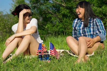 Two smiling asian woman with USA flags, celebration of patriotic american national holiday 4th of july independence day, festive picnic on lawn in parkの写真素材