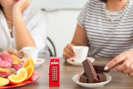 Traditional afternoon tea of british ceremony with such symbol of britishness as toy telephone box, defocused faceless women on background, front focusの写真素材