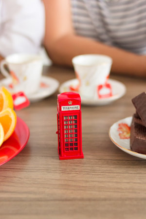 Traditional afternoon tea of british ceremony with such symbol of britishness as toy telephone box, defocused faceless women on background, front focus, vertical imageの写真素材