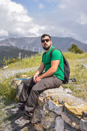 Hiking man taking a break sitting on a stone wall wearing sunglassesの写真素材