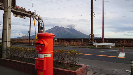 Kagoshima, Japan - 01.29.2020: Sakurajima releases smoke under a blue sky with a red Japanese postbox in the front on the pavement along an empty roadのeditorial素材