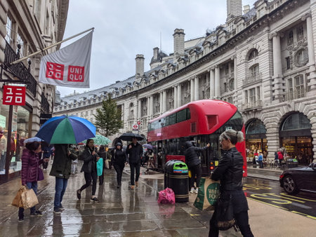 London, UK - 10.02.2021: Shoppers with umbrellas walking on Regent Street next to Uniqlo with cars and a double-decker bus disembarking passengers at a bus stop in the rainのeditorial素材