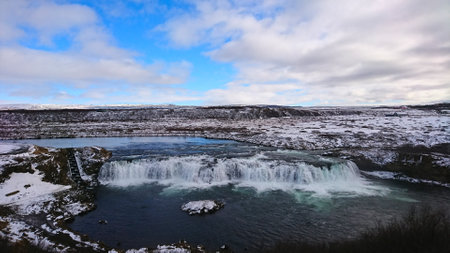 Faxafoss, Iceland - 3.24.2018: Faxi waterfall with dramatic water crashing into the lake through a snowy landscape and rugged terrain with snow mountains at the horizon under a blue sky with cloudsの写真素材