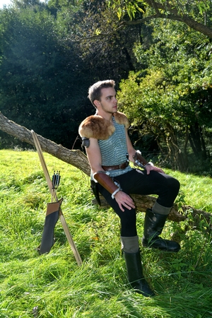 Young Medieval dressed man, sits on branch, arms on legs and looks aside, arrow and curve stands besidesの写真素材