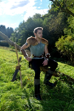 Young medieval archer with chain shirt sits on branch in the nature in the sunlight, drinking horn in the hand, arrow and curve stands besidesの写真素材