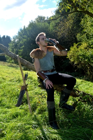 Young Medieval dressed man, sits on branch, drinks from drinking horn. Eyes open, arrow and curve stands besidesの写真素材