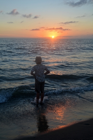 Small boy stands in the waves on the beach at the sundown, hands in the trouser pockets and looks in the waterの写真素材