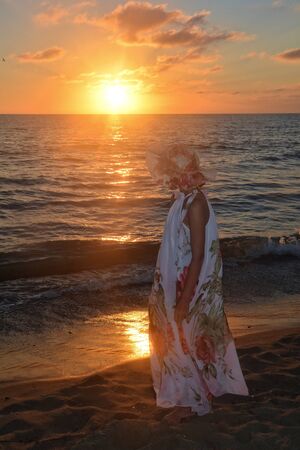 Girl in the long dress stands by the sea beach at the sundown, with look as the sunの写真素材