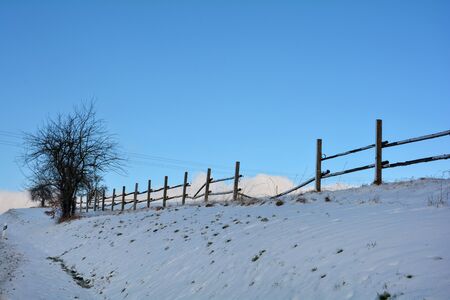 Wooden fence in winter with snow, blue sky and a tree on the leftの写真素材