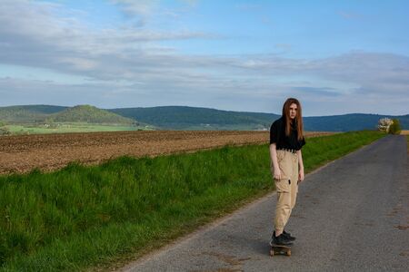 Young girl rides a skateboard on a country road in green nature, with blue sky and many copy spaceの写真素材