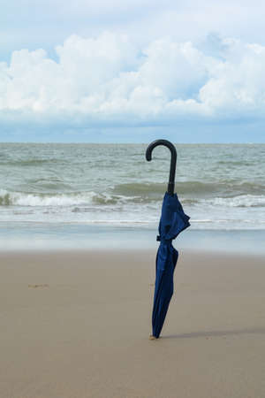 A closed umbrella is stuck in the sand on the beach in front of the ocean with waves and copy space