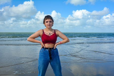Young girl in jeans and a short top stands  on the sandy beach in front of the sea with a blue sky and many cloudsの写真素材