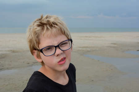 A boy stands with a black T-shirt on the beach, with a facial grimace, grains of sand on his glasses, at low tide in front of the North Sea coastの写真素材